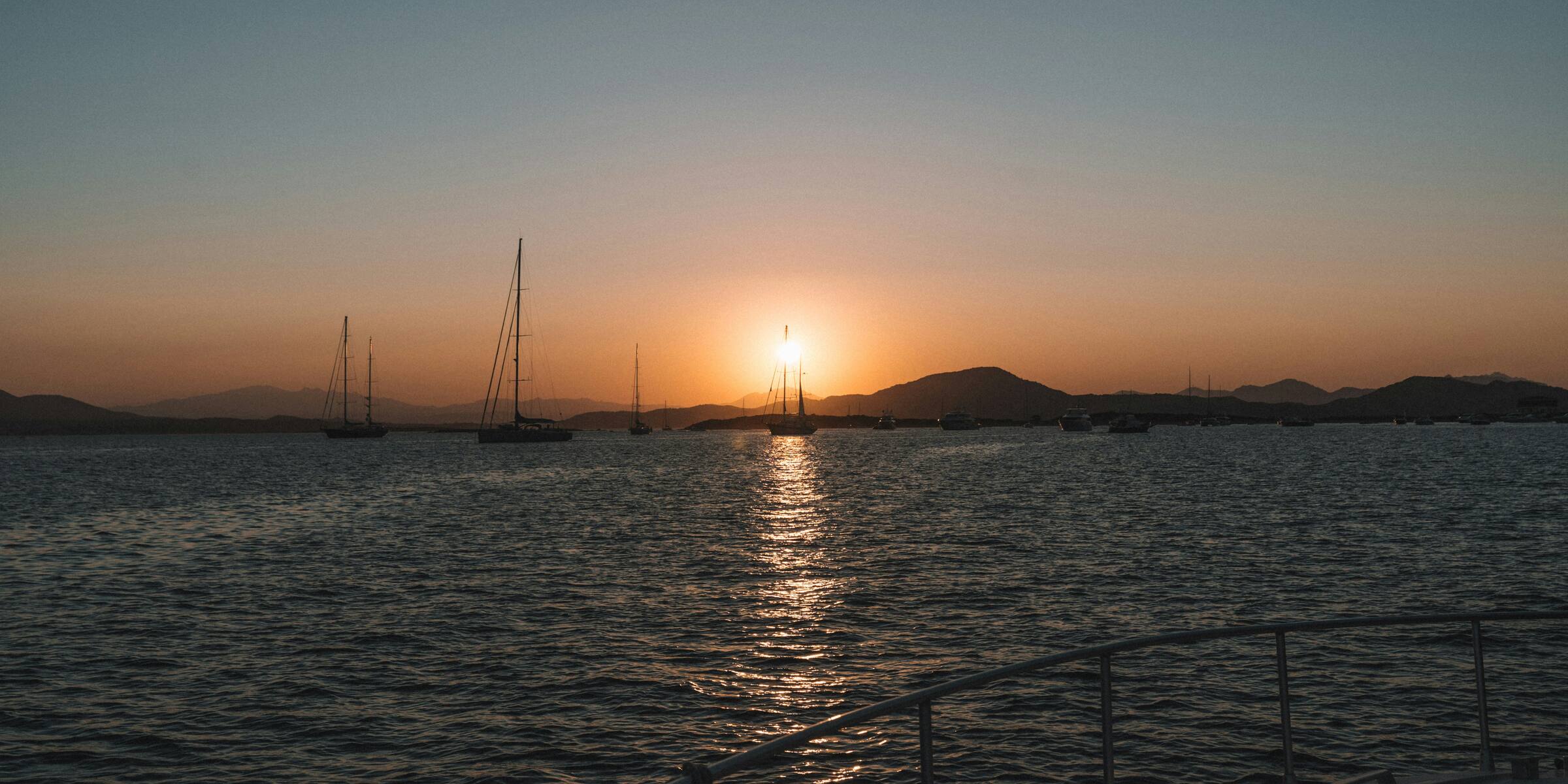 Blick über die Reling einer Yacht auf das Wasser bei Sonnenuntergang, am Horizont ankernde Segelboote vor einer Hügelkette.
