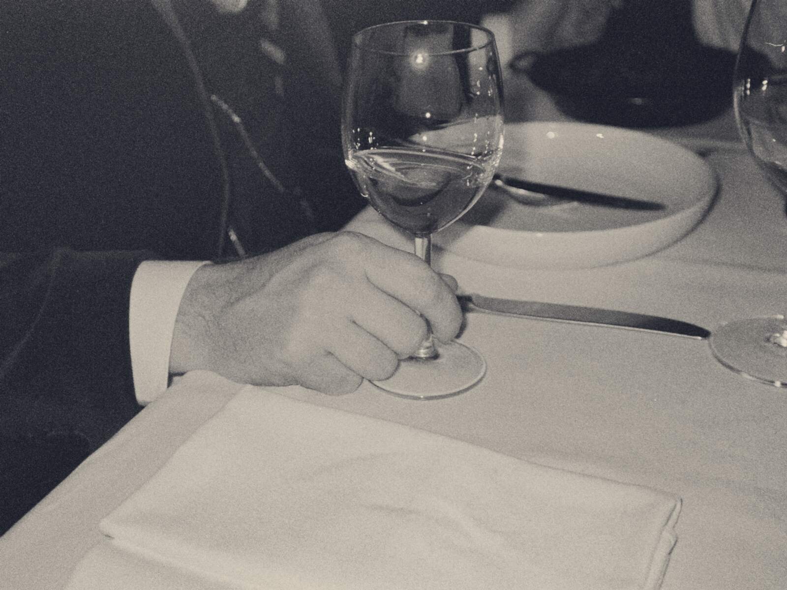 Eine Hand im Anzugärmel an einem Weinglas auf weiß eingedecktem Tisch, gegenüber ein zweites Glas.